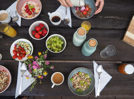 Un tavolo per la colazione con ciotole di cereali, fragole, uva, verdure a fette, bevande, tazze, posate, tovaglioli e un piccolo vaso di fiori. Una mano versa il latte in una ciotola.