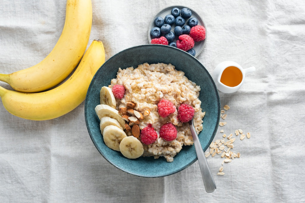Una ciotola di porridge con fette di banana, lamponi e noci, accanto due banane, un piatto di mirtilli e lamponi, una piccola brocca di miele e fiocchi d'avena sparsi su un panno.