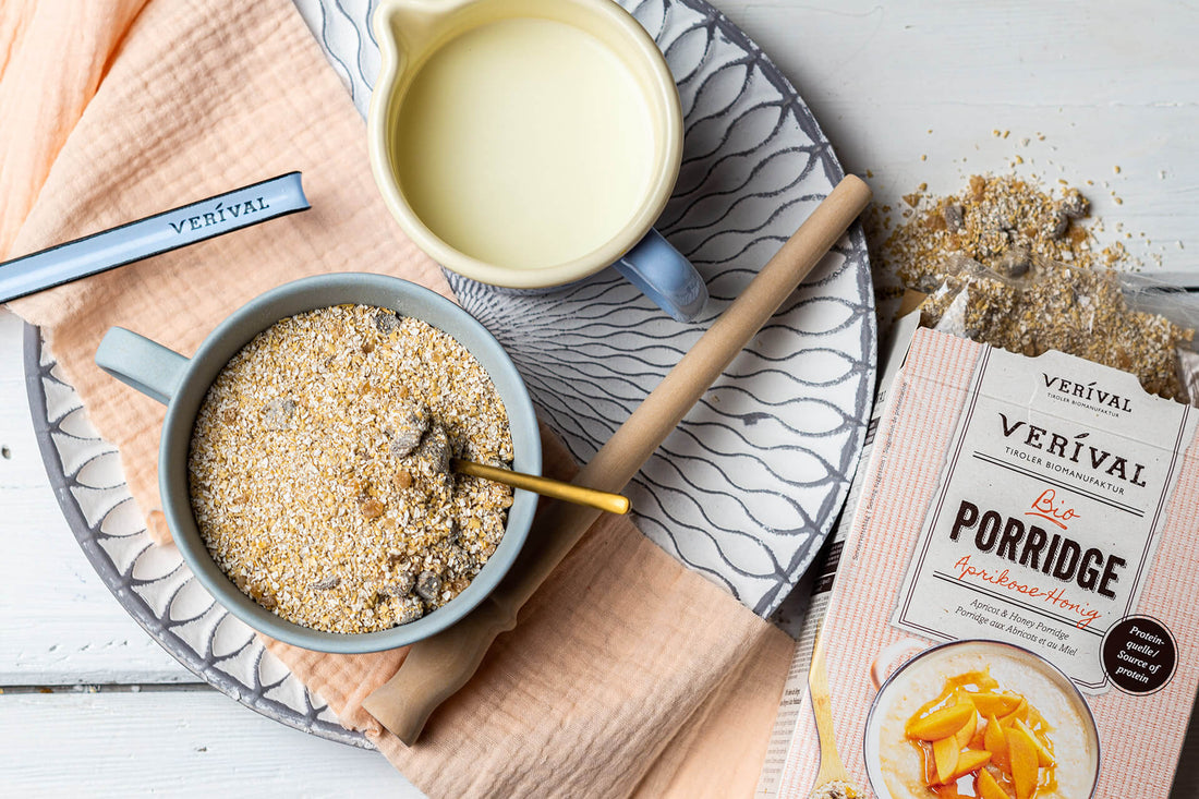 Una ciotola di porridge con un cucchiaio, una brocca di latte, una confezione di porridge Verival con albicocche e miele e un po' di porridge rovesciato su un vassoio con un tovagliolo.