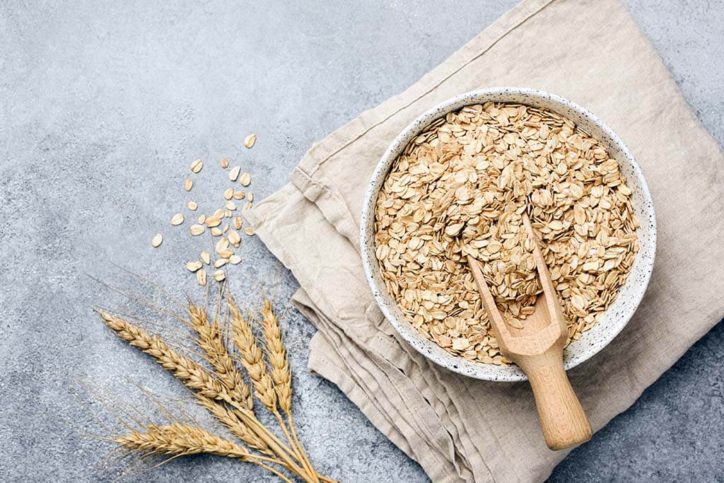 Una ciotola di fiocchi d'avena e un cucchiaio di legno sono appoggiati su un panno beige, accanto a spighe di grano e avena sparsi su una superficie grigia.