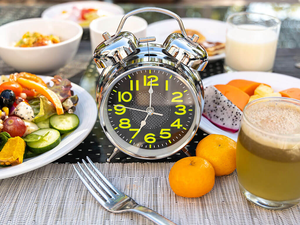 Una sveglia analogica argentata è posta su un tavolo tra vari prodotti per la colazione, tra cui insalata, frutta, succo di frutta e un bicchiere di latte.