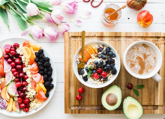 Una scena di colazione con una ciotola di frutta e cereali, un piatto con vari frutti freschi, un avocado, una tazza di caffè, miele, tulipani e una pesca su una superficie di legno.