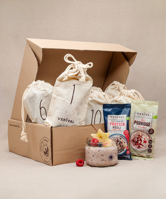 Ein Karton präsentiert drei Adventskalender-Säckchen von Verival Bio sowie zwei Packungen der Verival-Produkte Protein Müsli und Porridge. Im Vordergrund steht ein Glas palmölfreies Müsli, garniert mit Fruchtscheiben und Himbeeren.