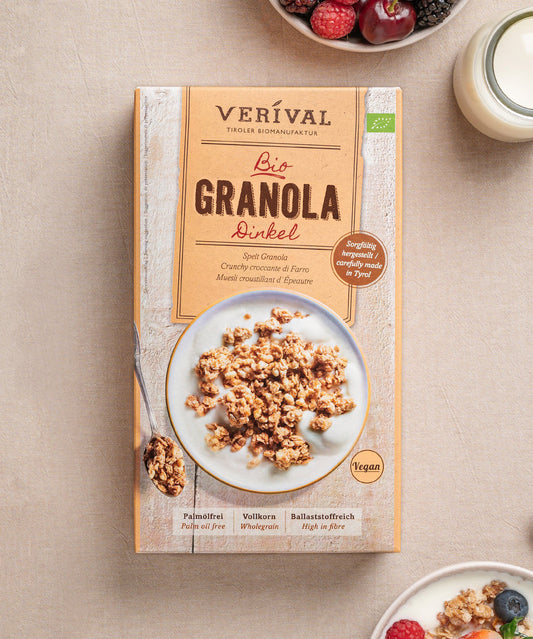 Auf dem Tisch steht eine Packung Dinkel-Granola von Verival Bio, ideal für ein Knusper-Frühstück. Daneben wartet eine Granola-Bowl mit Beeren und einem Glas Milch. Die Verpackung betont die vegane Eigenschaft, die Vollkornvorteile und den Verzicht auf Palmöl.