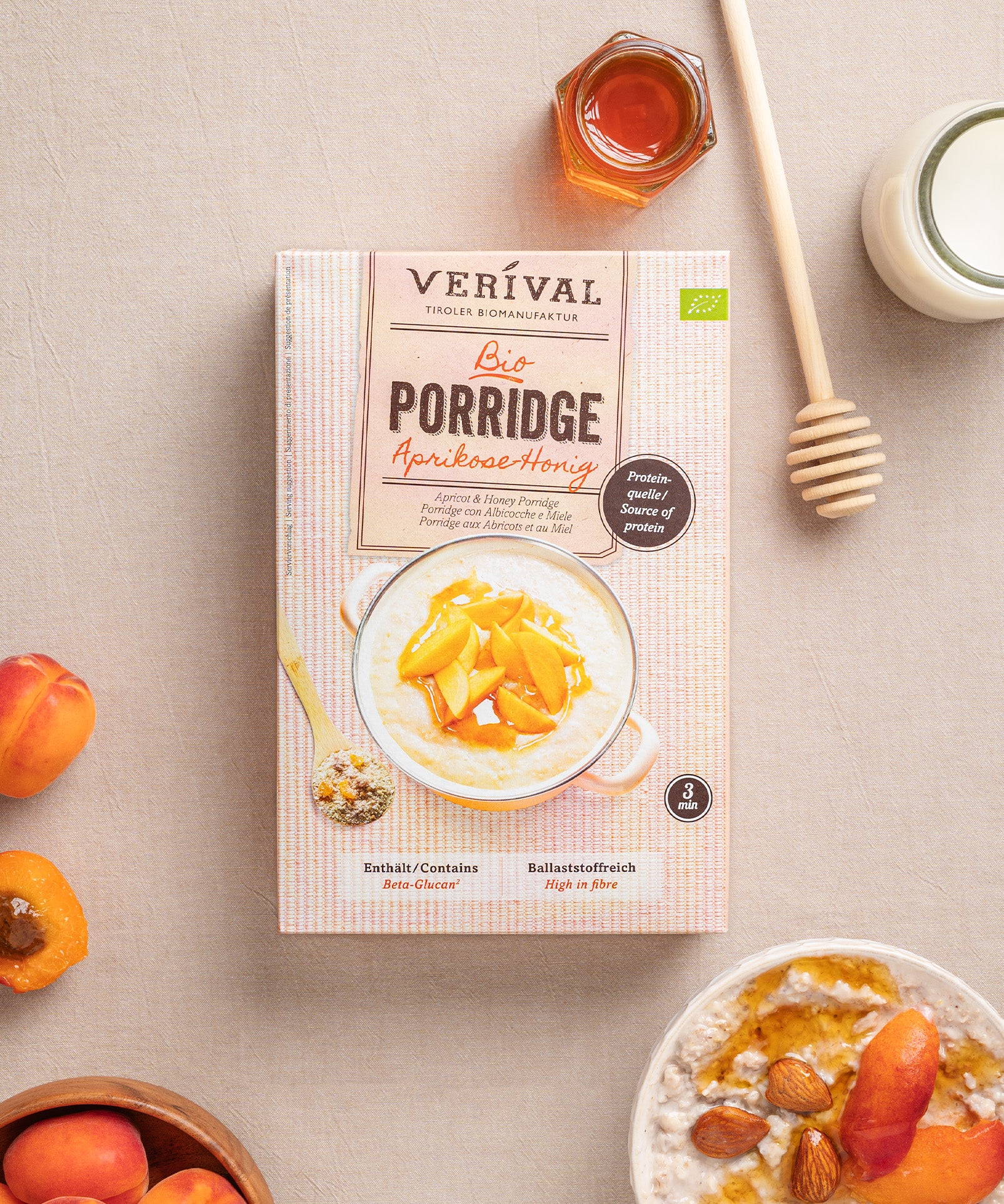 Eine Packung Aprikosen-Honig Porridge von Verival Bio  steht mit frischen Aprikosen, Mandeln, einem Honiglöffel, einem Glas Honig und Milch auf dem Tisch.