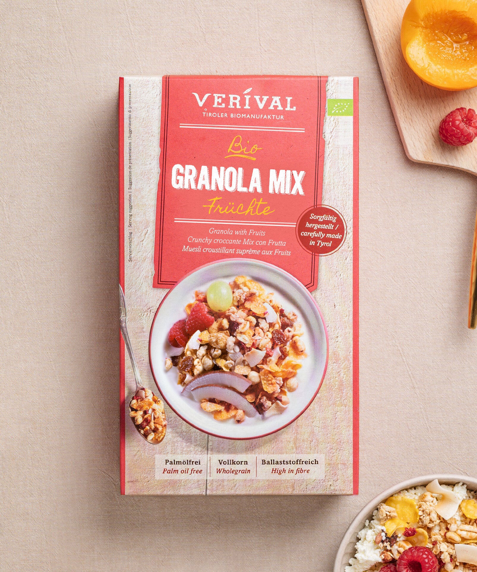 Eine Schachtel Granola Mix Früchte von Verival Bio   steht auf einem beigen Untergrund. Darin befindet sich eine farbenfrohe Schale mit Granola, Nüssen und Trockenfrüchten. Daneben steht eine weiße Schale mit proteinreichem Granola, umgeben von verstreuten frischen Früchten.