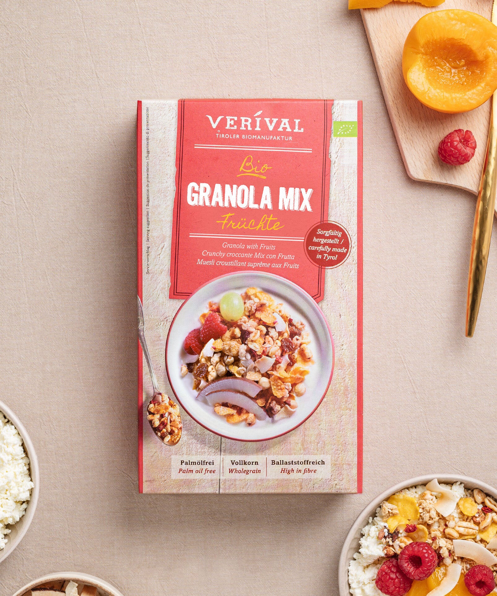 Auf einem Tisch steht eine Packung Granola Mix Früchte  von Verival Bio, daneben eine Schüssel mit dem fruchtigen und ballaststoffreichem Granola. Daneben liegen Pfirsichscheiben, Himbeeren und ein Holzlöffel und schaffen damit eine einladende Frühstücksszenerie.
