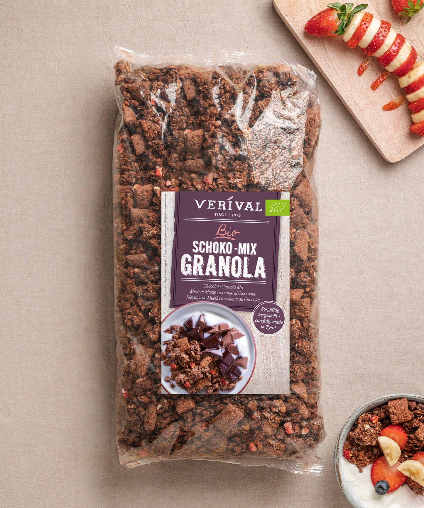 Eine große Packung Bio Schoko Mix Granola von Verival Bio GmbH steht auf beige, daneben eine Schale Joghurt mit Obst sowie ein Holztablett mit Apfel- und Erdbeerscheiben.