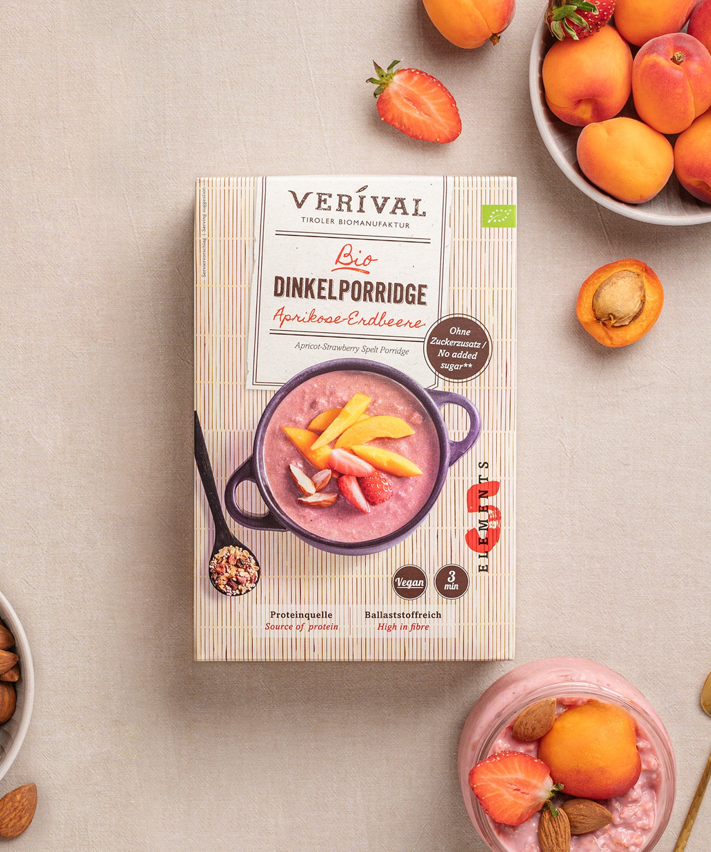 Eine Packung Aprikosen-Erdbeer Dinkelporridge von Verival Bio steht auf dem Tisch, umgeben von Schalen mit Aprikosen, Erdbeeren, Mandeln und einem Glas des nährstoffreichen, ballaststoffreichen Porridges. Perfekt für ein vollwertiges TCM-Frühstück.