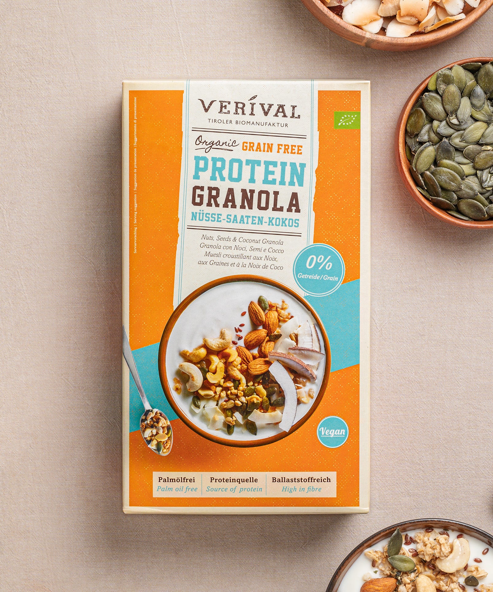 Eine Packung Grain Free Protein-Granola Nüsse-Saaten-Kokos von Verival Bio, reich an pflanzlichen Proteinen, steht auf dem Tisch, umgeben von Schalen mit verschiedenen Nüssen und Samen. Ideal für alle, die ein Low-Carb- oder Sport-Granola suchen, um Energie für den Tag zu tanken.