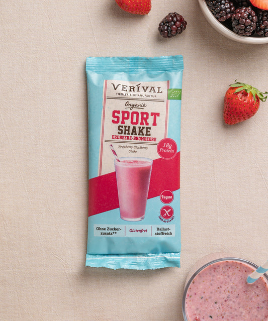 Eine Einzelportionspackung vom Sport Shake Erdbeere-Brombeere von Verival Bio ist umgeben von Erdbeeren, Brombeeren und dem Shake in einem Glas auf einem beigen Untergrund.