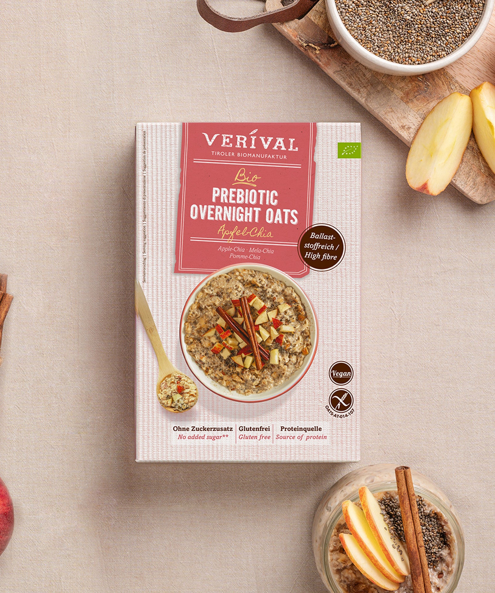 Die Prebiotic Overnight Oats Apfel-Chia der Verival Bio GmbH enthalten Äpfel und Chia sowie ballaststoffreiche, vegane, glutenfreie Haferflocken ohne Zuckerzusatz. Das Produkt präsentiert sich zwischen Haferflocken und Apfelscheiben auf einem beigen Untergrund.