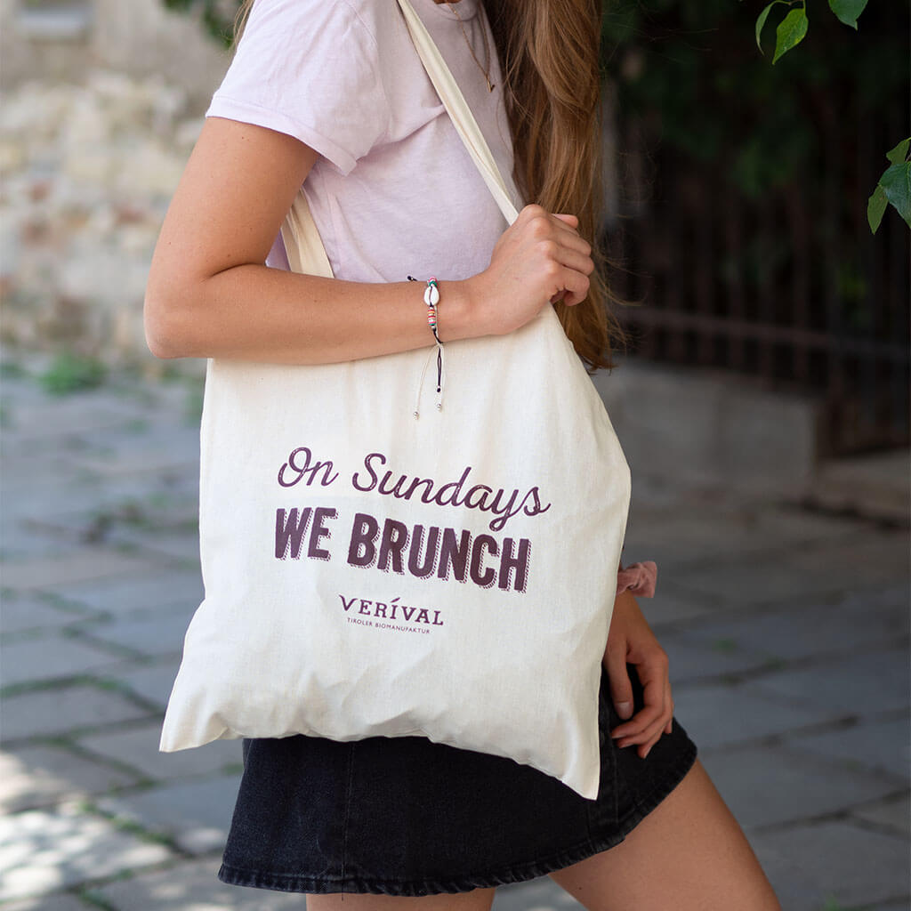 Eine Person in blassrosa T-Shirt und einen dunklen Rock hält eine Verival Bio-Stofftasche mit dem Schriftzug „On Sundays We Brunch“. Sie steht auf einem Kopfsteinpflasterweg im Schatten grüner Blätter und ist bereit für einen umweltfreundlichen Einkauf.
