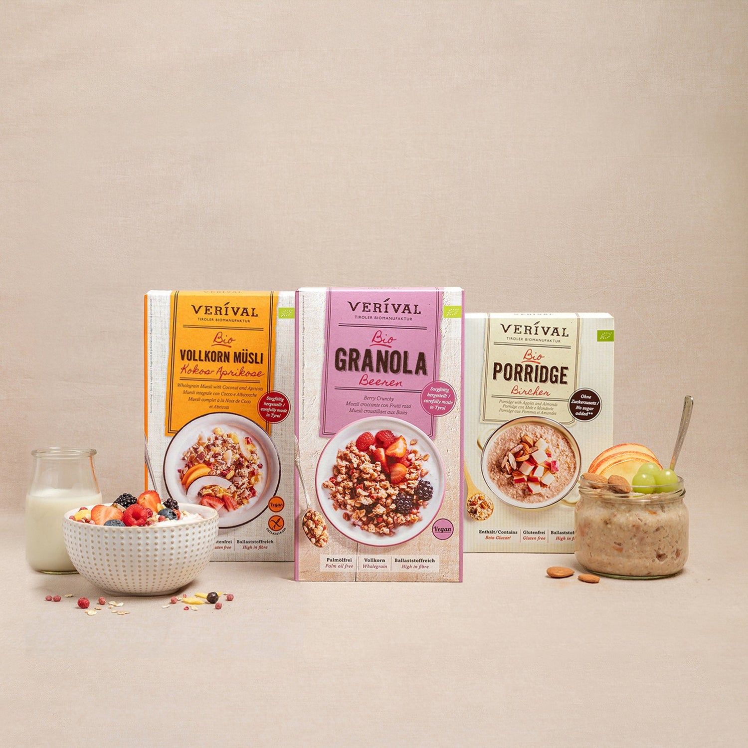 Tre scatole di cereali Verival: muesli integrale, granola ai frutti di bosco e porridge. Nella foto, una ciotola di muesli, una ciotola di porridge, un bicchiere di latte e frutta su sfondo beige.