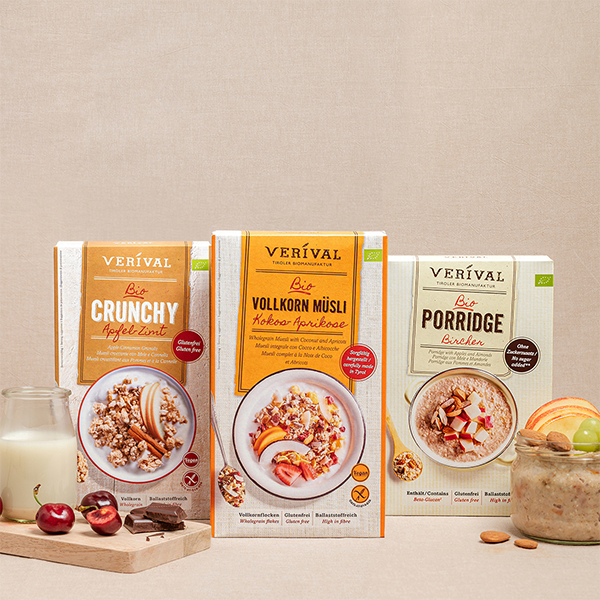 Tre scatole di cereali etichettate Crunchy, Wholegrain Muesli e Porridge sono presentate con un bicchiere, ciliegie e mandorle su uno sfondo beige.