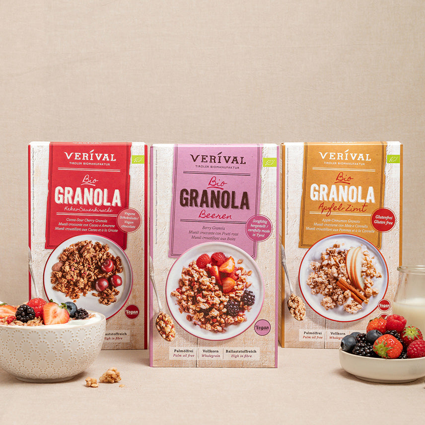 Tre scatole di granola Verival in vari gusti sono sistemate in verticale su una superficie beige, con una ciotola di granola e frutti di bosco misti di fronte.