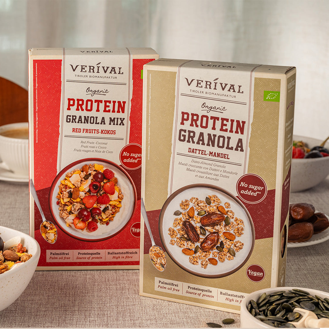 Su un tavolo con ciotole di granola e ingredienti ci sono due scatole di granola proteica biologica Verival, una con frutti rossi e cocco e una con datteri e mandorle.