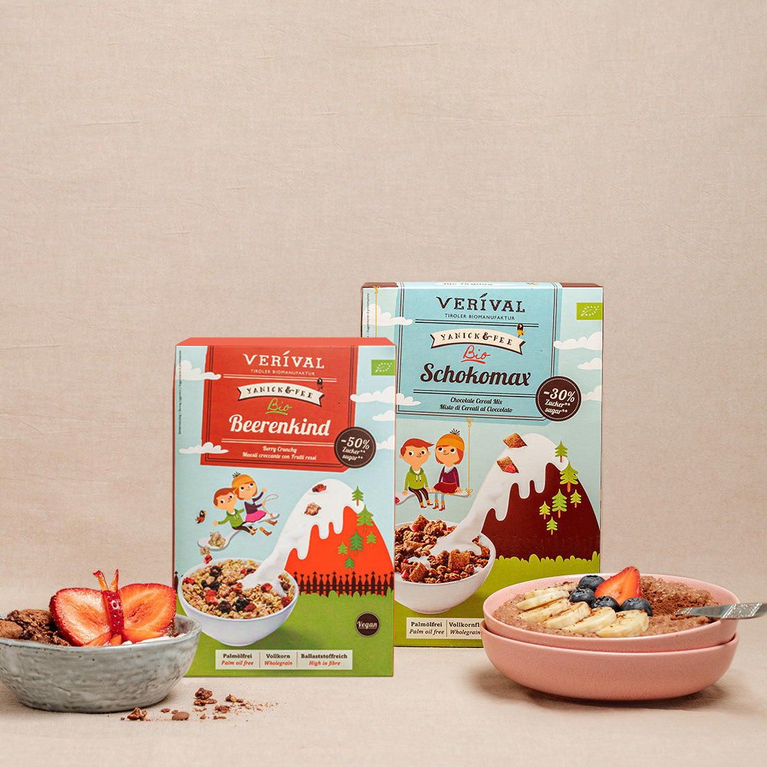 Due confezioni di muesli Verival, Beerenkind e Schokomax, sono posizionate in verticale accanto a una ciotola di muesli con fette di fragole e banana su uno sfondo beige.