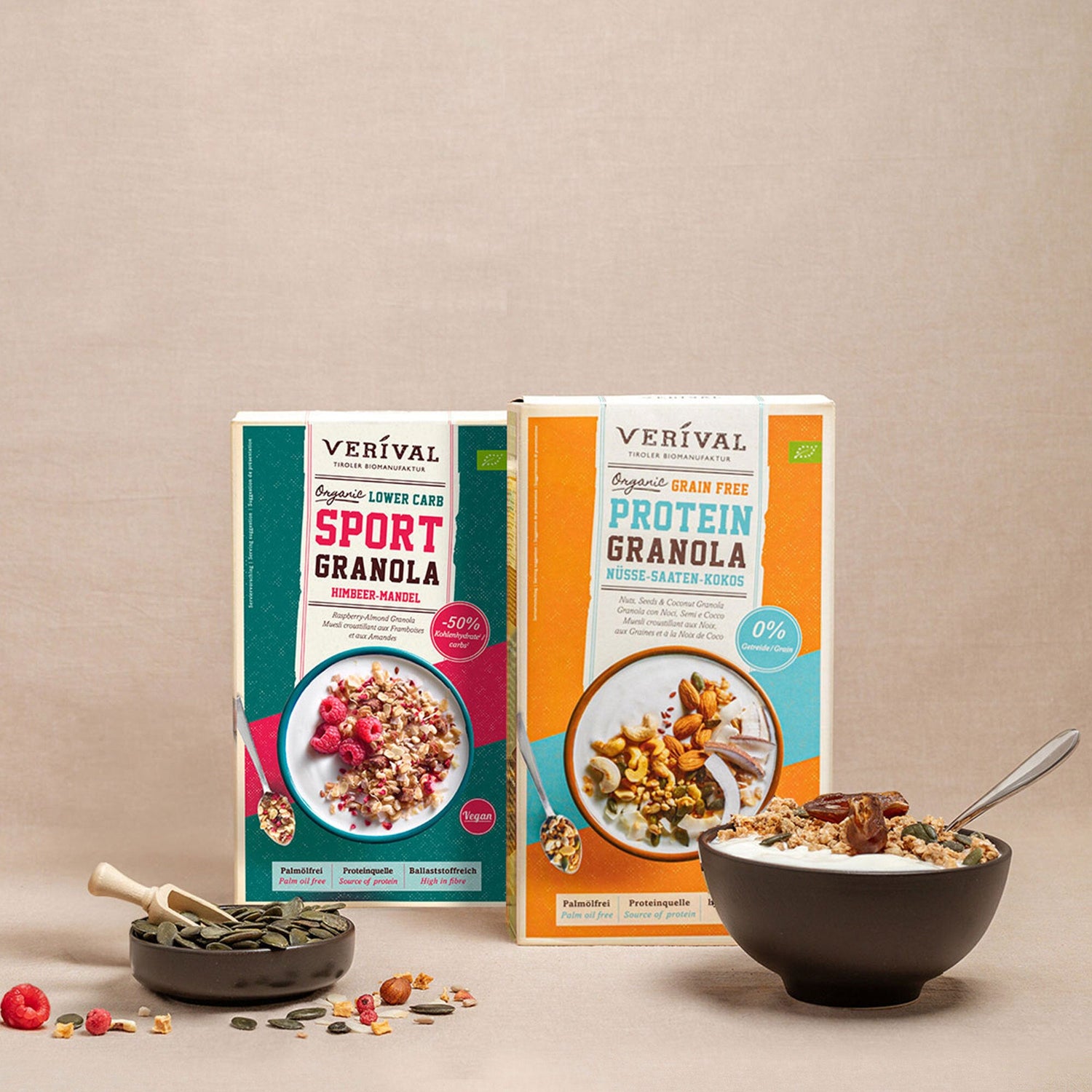 Due scatole di granola Verival (Sport Granola e Protein Granola) sono posizionate dietro una ciotola di granola con un cucchiaio e una piccola ciotola di semi su uno sfondo neutro.