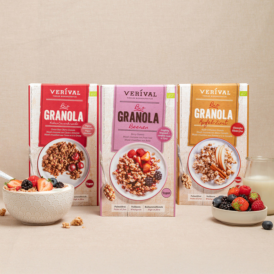 Tre scatole di muesli integrale Verival in diversi gusti vengono presentate con una ciotola di muesli, un bicchiere di latte e vari frutti di bosco su uno sfondo beige.