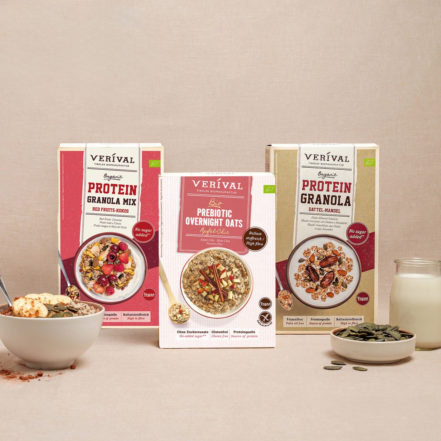 Tre confezioni di cereali per la colazione Verival, tra cui Protein Granola Mix, Prebiotic Overnight Oats e Protein Granola. Davanti, una ciotola di muesli, un cucchiaio, frutta secca e un bicchiere di latte.