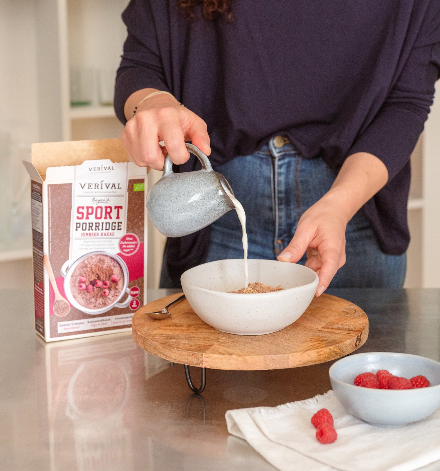 Una persona versa il latte in una ciotola di porridge accanto a una scatola con l'etichetta Sport Porridge e a una piccola ciotola di lamponi su un vassoio di legno.
