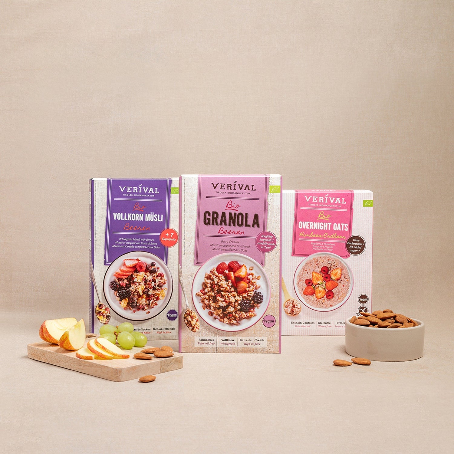 Tre confezioni di muesli Verival (muesli integrale, granola, fiocchi d'avena durante la notte) vengono presentate con una ciotola di mandorle e un tagliere con fette di mela e uva.