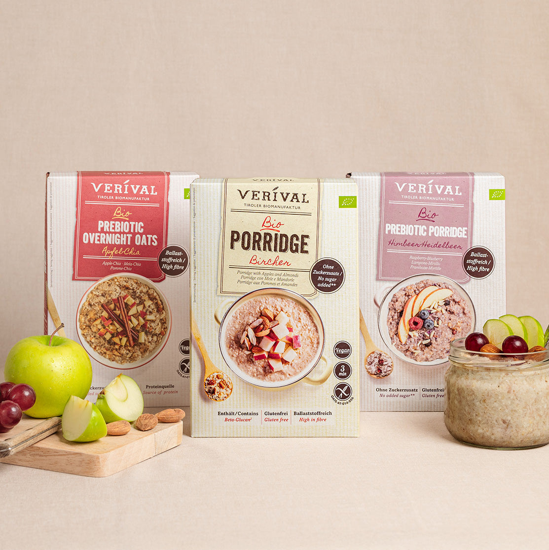 Tre confezioni di cereali per la colazione Verival (Prebiotic Overnight Oats, Organic Porridge e Prebiotic Porridge) presentate con una ciotola di porridge, fette di mela e uva davanti.