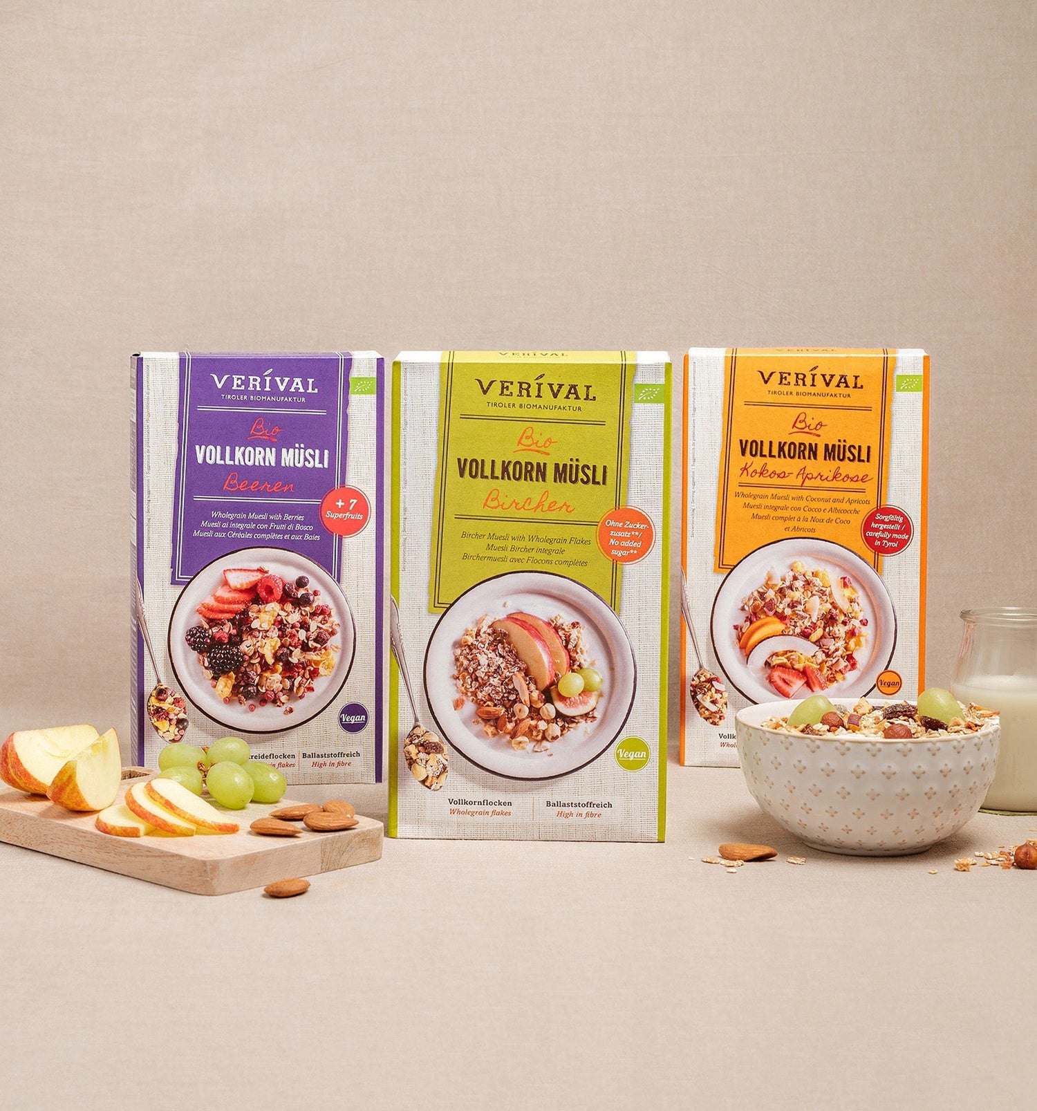 Tre scatole di muesli integrale biologico Verival sono posizionate in verticale, davanti a loro una ciotola di muesli, fette di mela, uva, un cucchiaio e un bicchiere di latte.