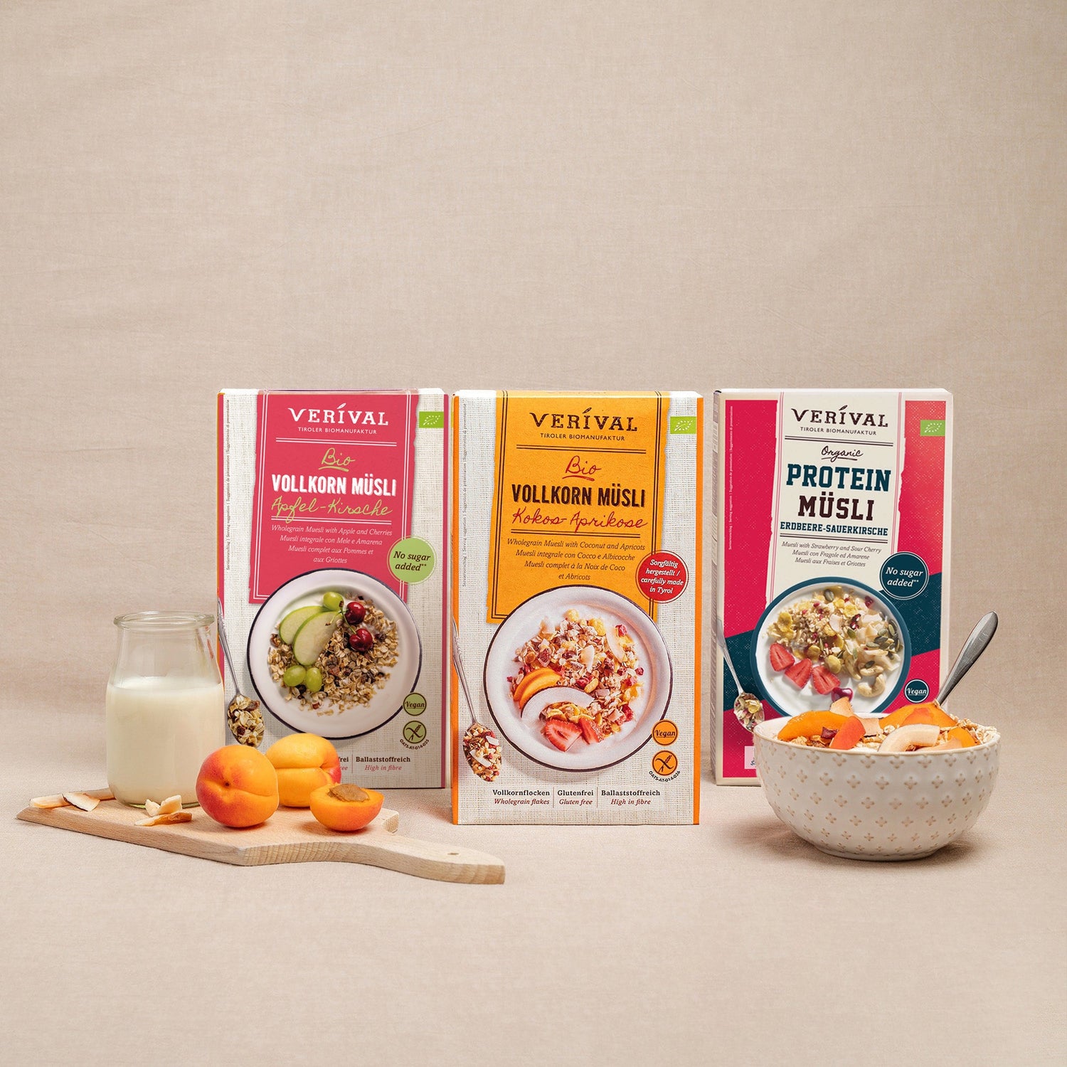 Tre scatole di muesli integrale biologico Verival in vari gusti sono sistemate in verticale su una superficie beige, accanto a una ciotola di muesli, fette di mela, uva e un bicchiere di latte.