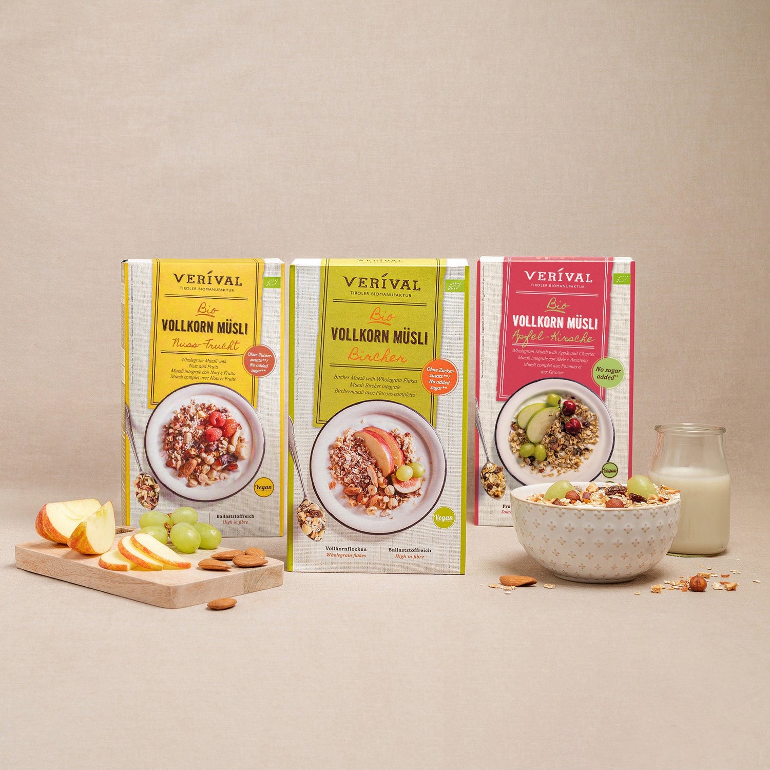 Tre scatole di muesli integrale Verival in vari gusti sono presentate in verticale, con una ciotola di muesli, un bicchiere di latte, fette di mela e uva davanti.