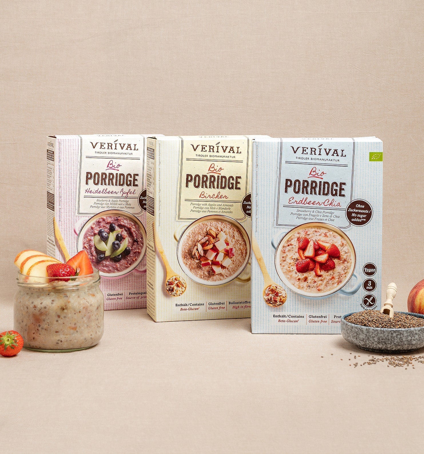 Tre scatole di porridge biologico Verival (nelle varietà mela, Bircher e chia) vengono presentate con un bicchiere di porridge, frutta fresca, semi di chia e una mela su uno sfondo beige.