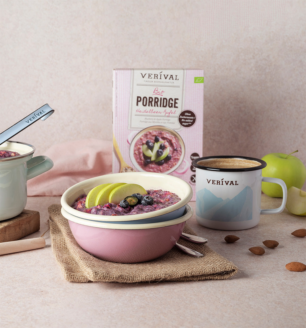 Tre scatole di porridge biologico Verival in diversi gusti sono posizionate accanto a un barattolo di porridge pronto, fragole, fette di mela e una ciotola di semi di chia.