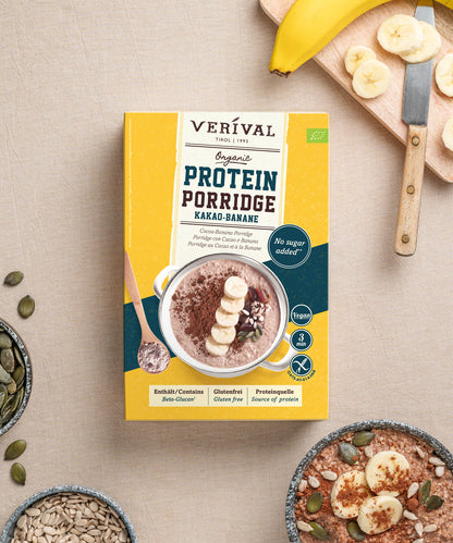 Eine Packung Verival Bio Bio Kakao-Banane Protein-Porridge steht auf einer beigen Unterlage, umgeben von einer in Scheiben geschnittenen Banane, Sonnenblumenkernen und einer Schüssel mit dem zubereiteten, proteinreichen Frühstück.