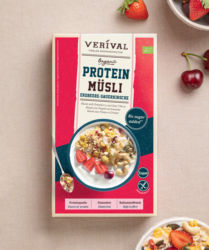 Eine Packung Protein Müsli Erdbeere-Sauerkirsche der Verival Bio GmbH steht auf einem hellen Untergrund mit frischen Kirschen und einer Schale Sport Müsli. Diese pflanzliche Proteinquelle wird ohne Zuckerzusatz hergestellt und sorgt für einen natürlich leckeren Start in den Tag.