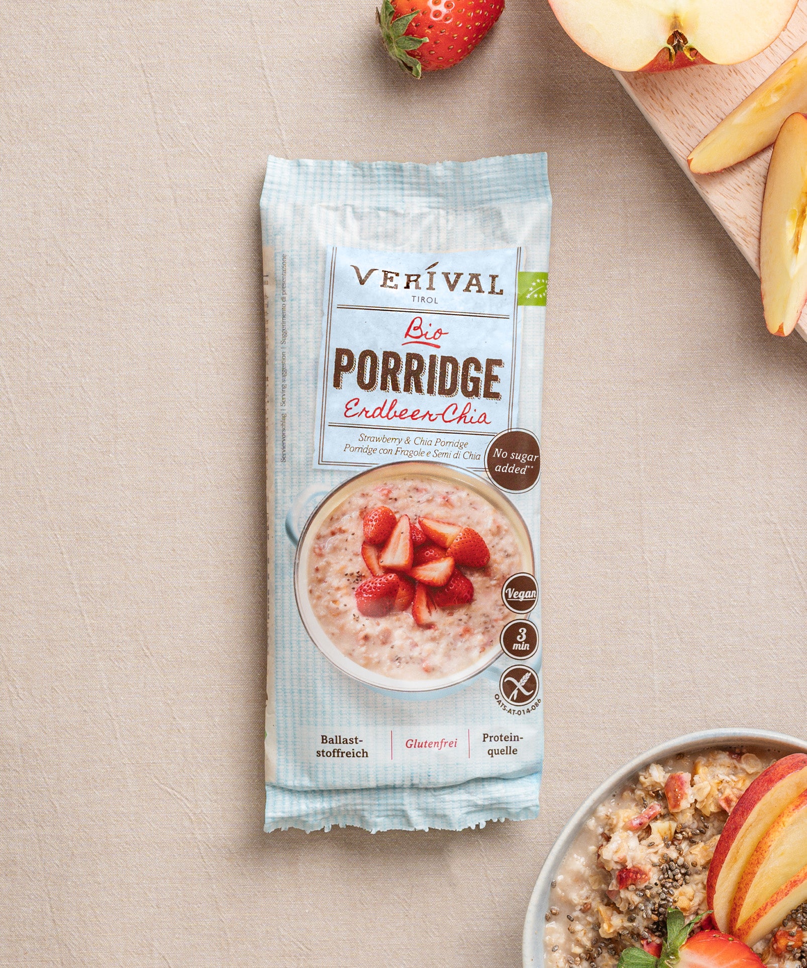 Eine Packung Erdbeer-Chia-Porridge der Verival Bio GmbH steht auf einer beigen Oberfläche, umgeben von einer pflanzlichen Frühstücksszene mit einer Schüssel Porridge, Apfelscheiben, frischem Obst und einer Erdbeere.