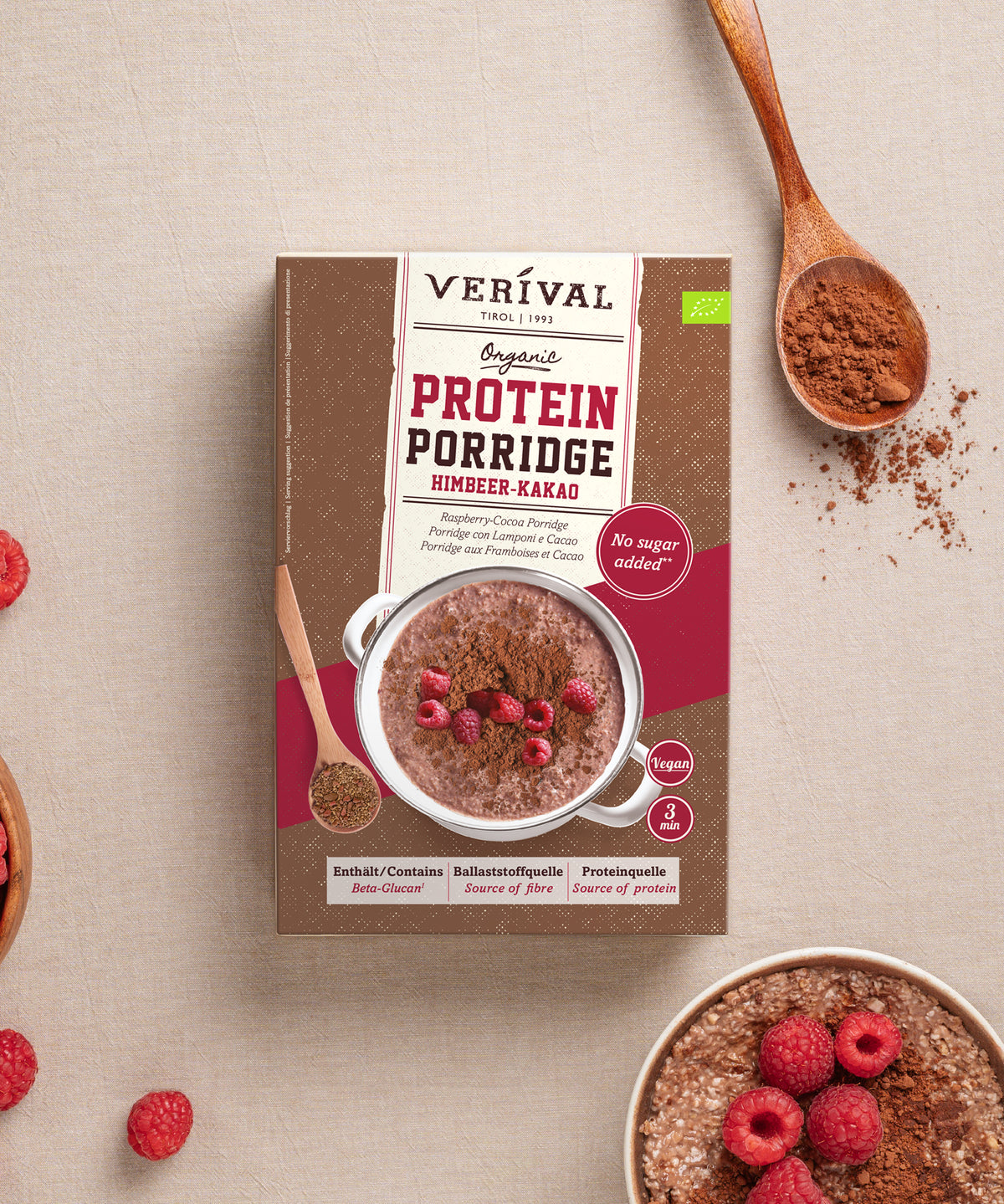 Eine Packung Bio Himbeer-Kakao Protein Porrige der Verival Bio GmbH, abgebildet zusammen mit einer Schüssel fertig zubereitetem Porridge, darauf verstreuten frischen Himbeeren und Löffeln mit trockenen Zutaten.