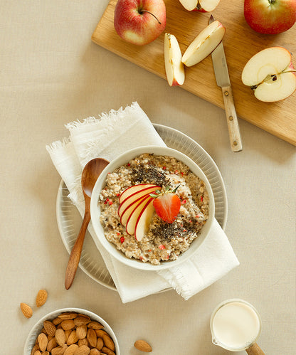 Auf einem Teller steht eine Schüssel mit Bio-Erdbeer-Chia-Porridge der Verival Bio GmbH, garniert mit Apfelscheiben, Chia-Samen und einer Erdbeere; daneben liegen Äpfel auf einem Schneidebrett mit einem Messer, Mandeln in einer Schüssel und ein Krug Milch.