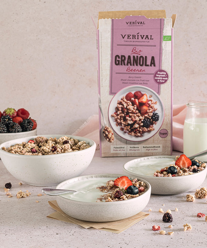 Auf einer hellen Unterlage stehen drei Schalen mit veganem Müsli, Milch und Beeren. Ein Glas Milch, eine Packung Bio-Beeren-Müsli von Verival Bio GmbH und eine kleine Schale mit gemischten Beeren runden dieses knusprige Frühstück ab.